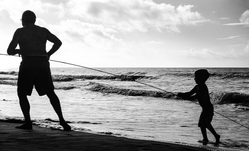 Silhouette of a kūpuna and a child fishing together on a Hawaiʻi beach at sunrise.